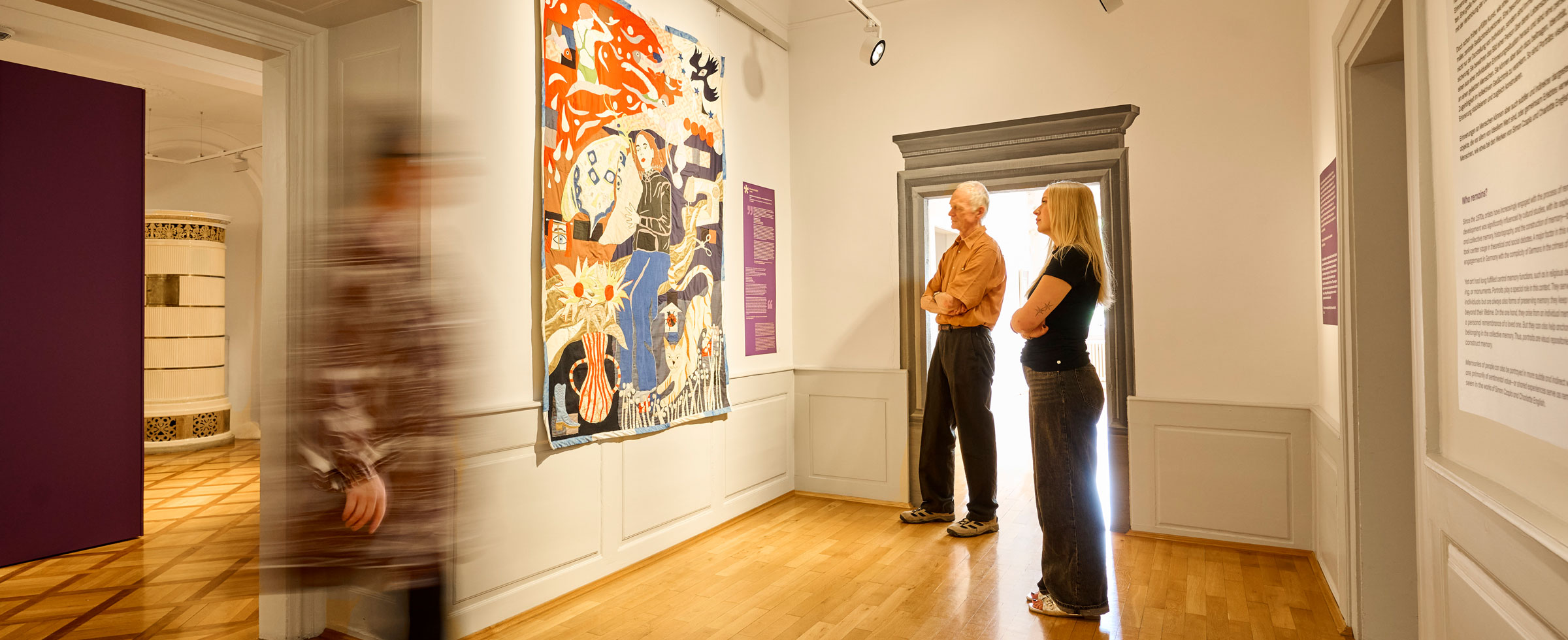 Ausstellungsraum der Wessenberg-Galerie Konstanz zur Ausstellung „Was bleibt. Erinnern und Vergessen in der Kunst“. Ein älterer Mann und eine junge Frau betrachten einen großen Wandbehang mit figürlichen Motiven, während im Vordergrund eine Person mit Bewegungsunschärfe vorbeigeht.